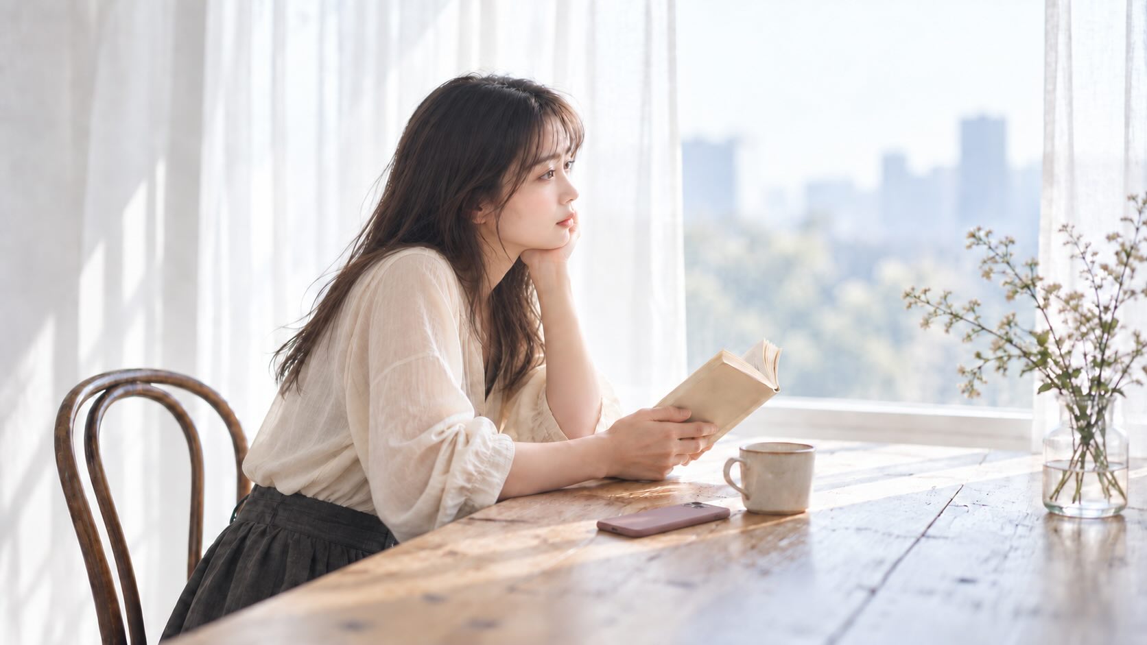 窓辺の木のテーブルで、文庫本を手に外を眺める女性