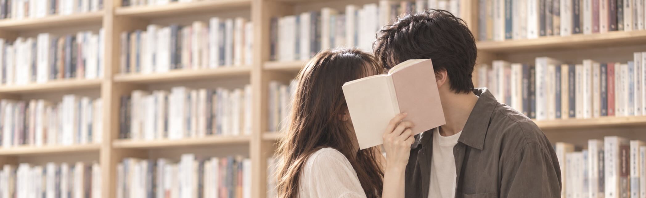 明るい本棚の前で、開いた本で顔を隠して寄り添う二人